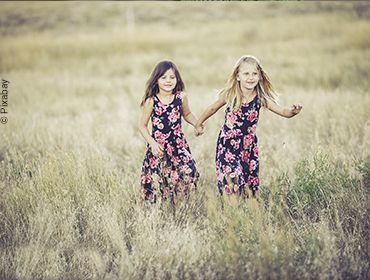 Zwei Mädchen laufen Händchenhaltend im Sommerkleid durch ein Kornfeld.