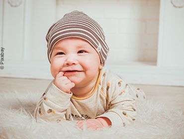 Lächelndes Baby liegt auf dem Bauch eines Teppichs mit erhobenem Kopf und einem Finger im Mund